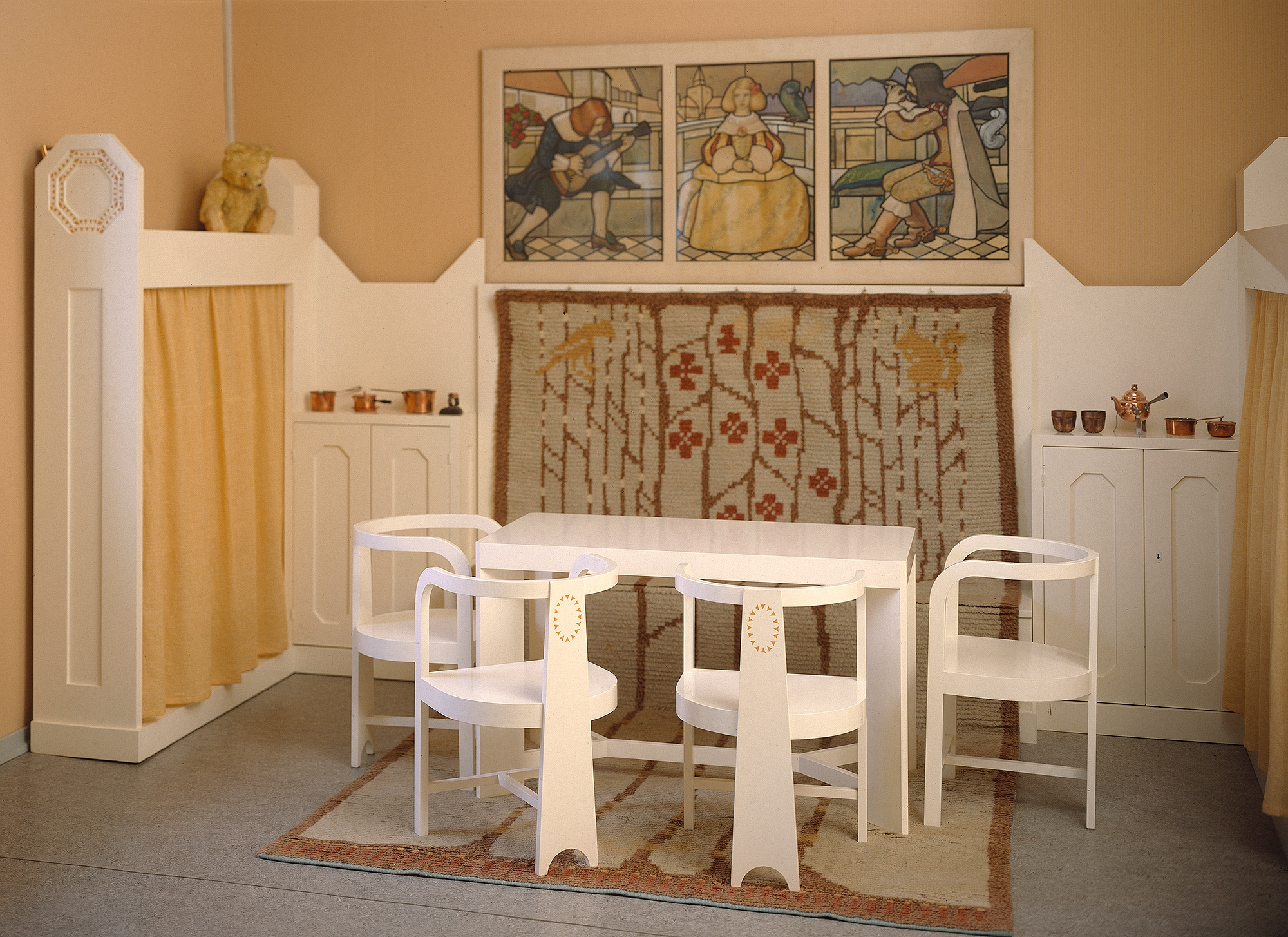 Keltaseinäisessa huoneessa valkoisia lapsille mitoitettuja puukalusteita ja seinällä kolmioisainen maalaus.