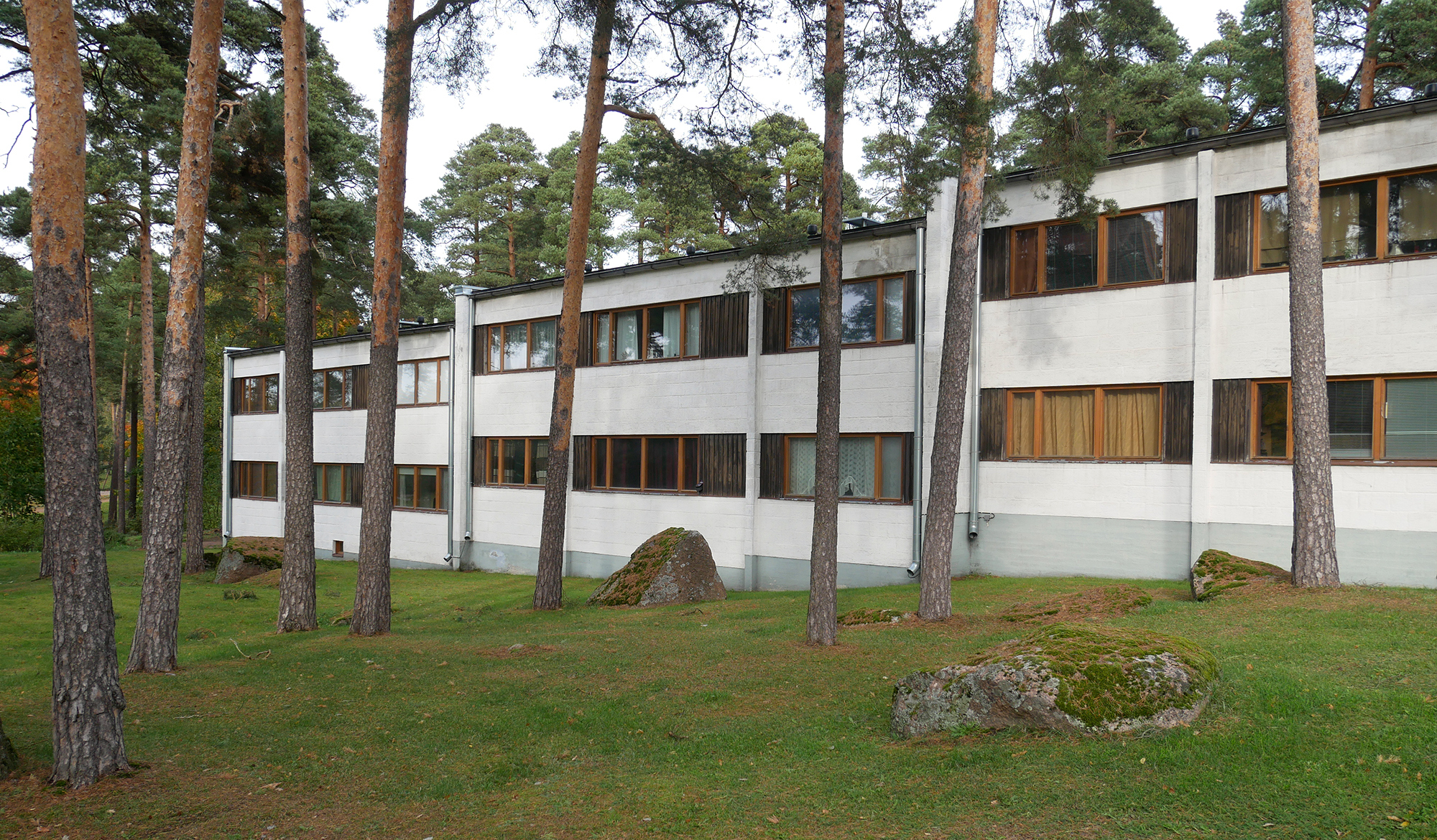 Kaksikerroksinen usean porrashuoneen kerrostalo kumpuilevassa mäntymetsässä. Vaalea rappauspinta ja nauhamaiset ikkunat korostavat rakennuksen pituutta.