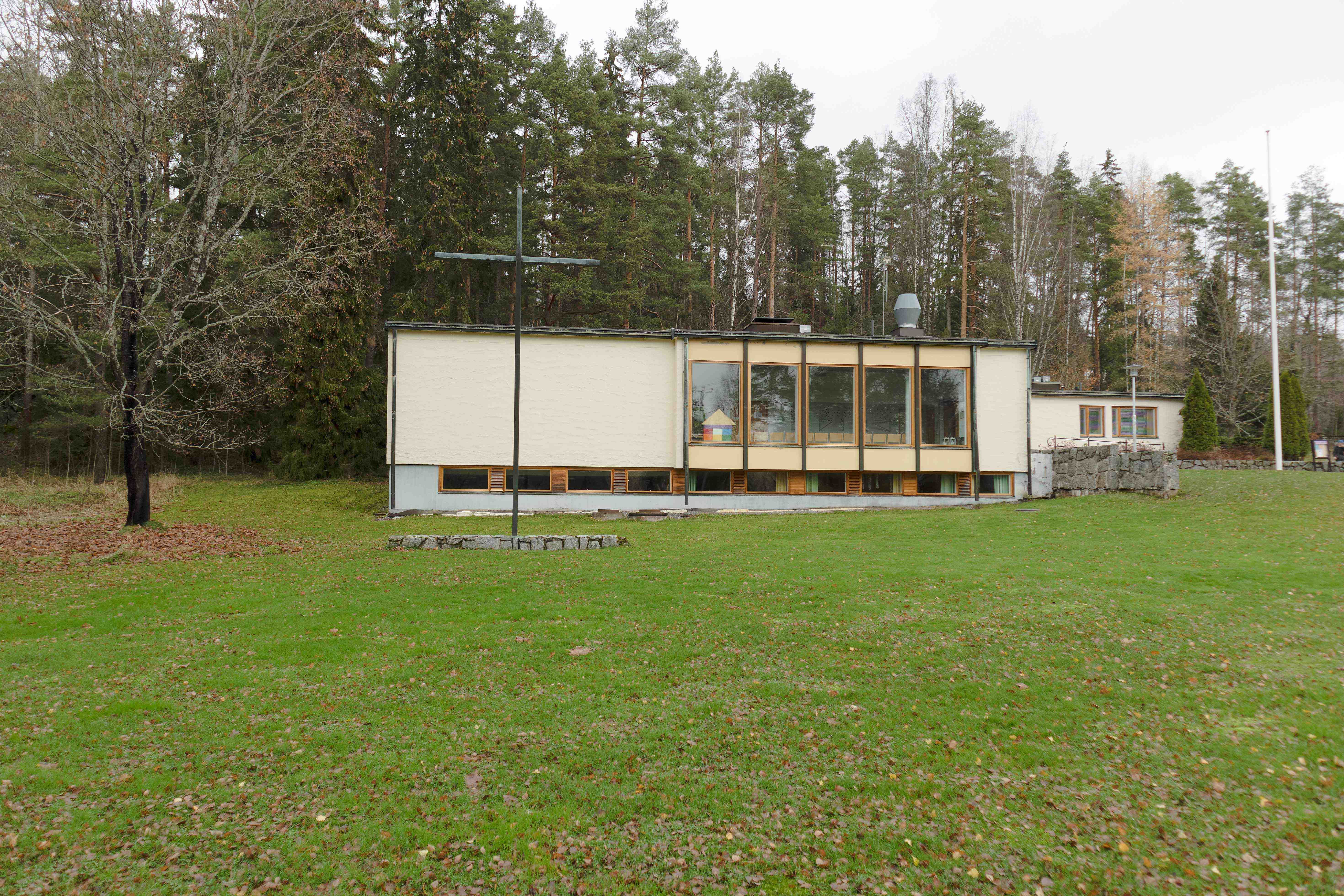 Yleiskuva nurmipintaisessa puistossa sijaitsevasta pienimittakaavaisesta modernista rakennuksesta. Rakennus on keltasävyinen, sen julkivivussa on suuri ikkunapinta ja rakennuksen vieressä on luonnonkivimuuri. Rakennuksen takana kasvaa täysikasvuinen metsä.