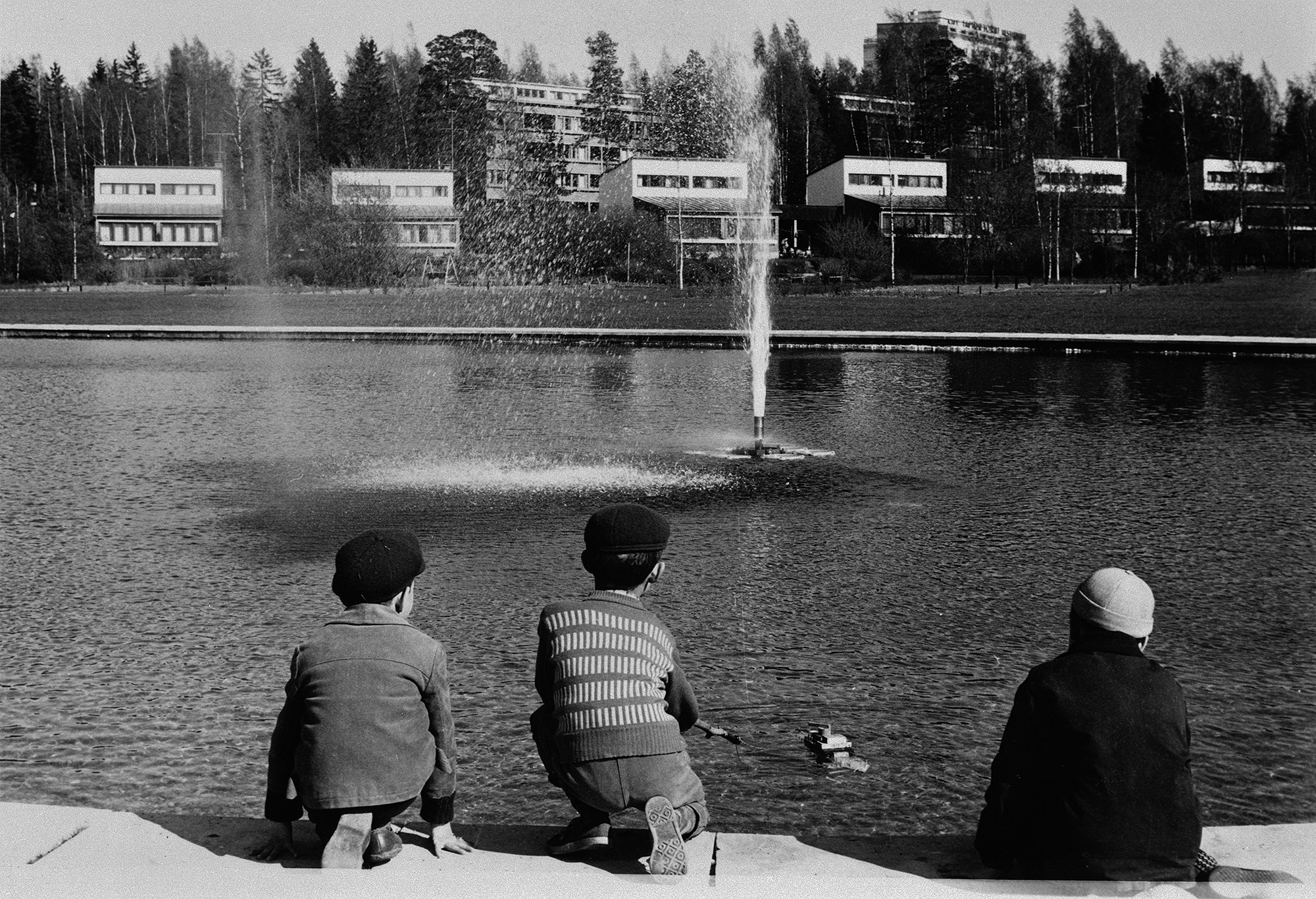 Mustavalkoisessa valokuvassa lapsia leikkimässä vesialtaan äärellä. Taustalla pientalorivi ja metsää.