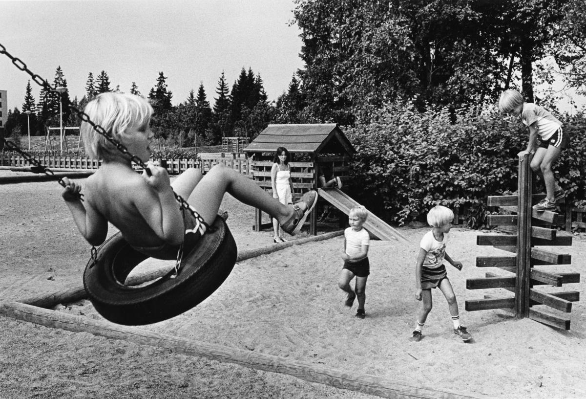 Yksi lapsi keinuu vauhdikkaasti rengaskeinussa, toinen kiipeää kiipeilytelineellä, kaksi muuta juoksee, aikuinen katsoo taustalla. Leikkikenttä on reunustettu pensailla, kentän pohja on hiekkaa ja leikkivälineet pääasiassa puisia.