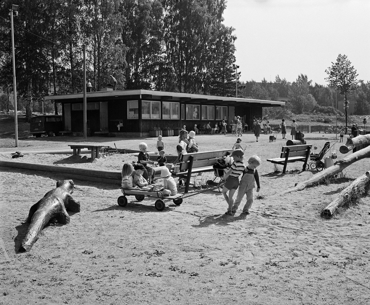 teksti: Lapset vetävät toisiaan kärryillä, leikkivät hiekkalaatikolla ja liikkuvat kesäisellä Puotinharjun leikkikentällä. Tehdasvalmistelisten leikkivälineiden lisäksi puistossa on mm. veistetty krokotiili kiinteänä leikkivälineenä.